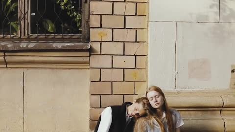 Women Leaning on Each Other While Sitting on a Sidewalk of a Street