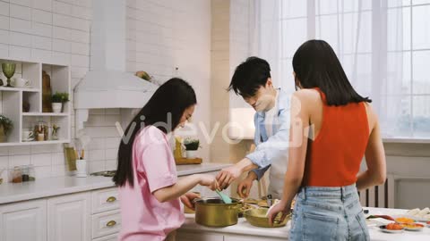 Three Japanese Friends Pouring Ingredients Into The Pan While Cooking, Talking And Laughing In The K