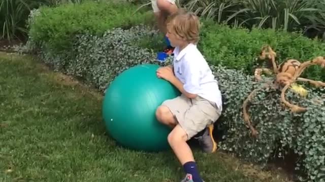 Boy in white jumps on green ball falls in grass