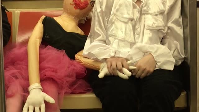 Girl dressed as a mime holds hands with a mannequin doll on subway train