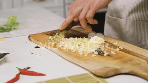 Close Up View Of Two Hands Chopping Garlic With A Knife On A Kitchen Board On The Countertop