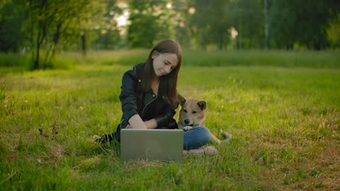 A woman in a park next to an open laptop is petting her dog