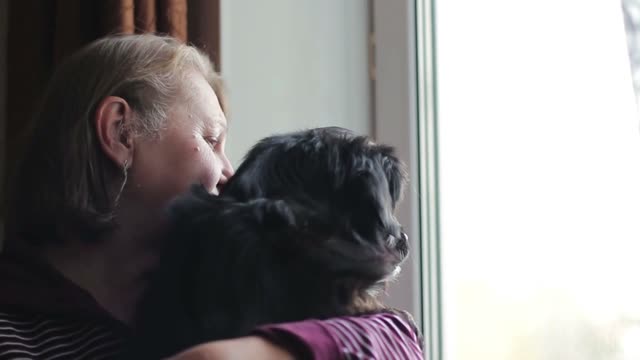 Aged woman holding a puppy in her arms and looking out the window together