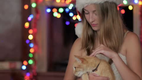 A smiling young attractive woman wearing a Santa hat petting an orange