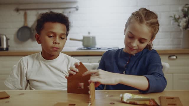 a boy watching a girl trying to make a model house made of cookie