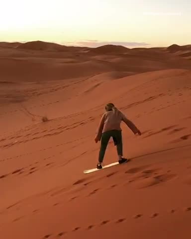 Guy rides white snowboard down sand dune and falls down