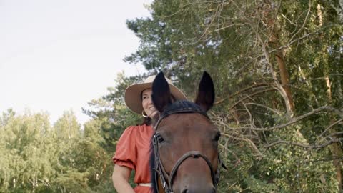 Smiling woman in hat sitting on brown horse on summer walk in forest