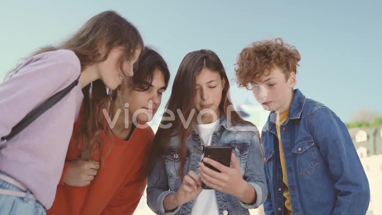 A Group Of Teenagers With Two Girls And Two Boys Watching Something On The Screen Of A Mobile Phone