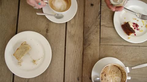 Overhead View Of Hands Lifting Cups Of Coffee And Cake Off Cafe Table