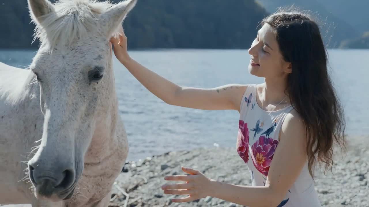 Pregnant woman petting white horse by the mountain lake at beautiful sunlight