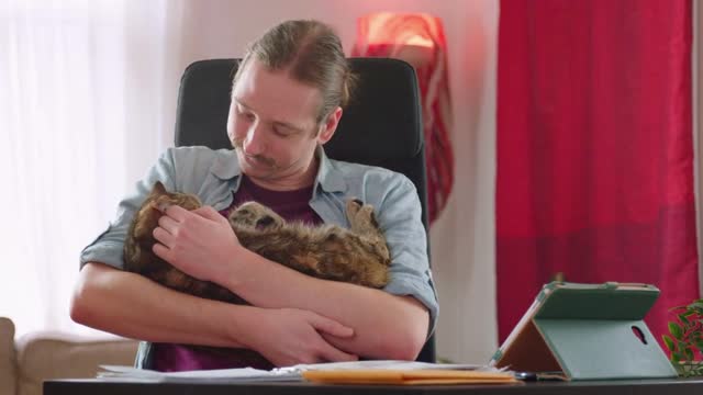 A caucasian man cuddles a cat affectionately while working from home - slow motion