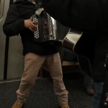 Guy on subway plays accordion and gives thumbs up to camera
