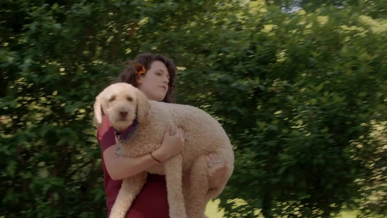 Young woman with flower in hair carries lazy dog across lush summer environment