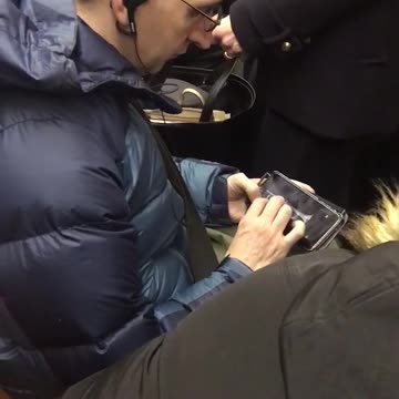 Man in blue jacket playing trumpet on phone sitting in crowded subway