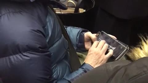 Man in blue jacket playing trumpet on phone sitting in crowded subway