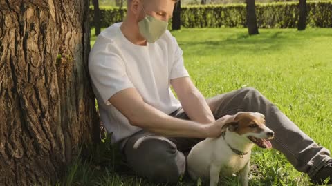 Handheld slowmo shot of young man in cloth face mask sitting on green grass under tree