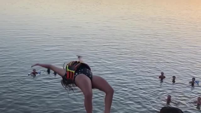 Girl in safety jacket does back flip off diving board into ocean