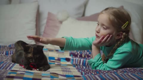 Adorable elementary age girl petting cute small cat with love while lying on bed