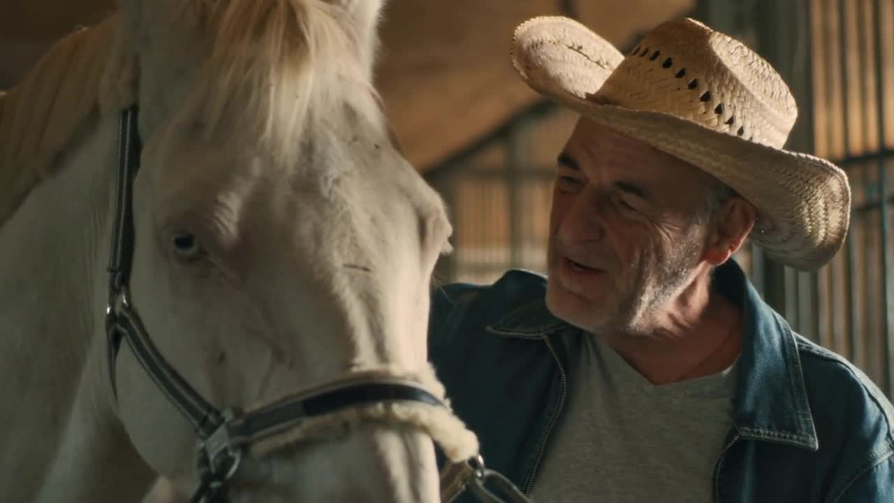 Positive senior man in straw hat embracing and kissing head of albino horse while