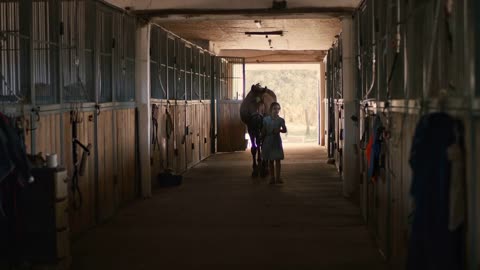 Little girl in dress walking in aisle in barn and leading chestnut horse