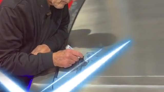 An old man signs his name on the hood of a car