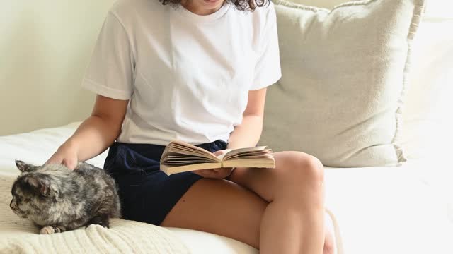 Woman Sitting on the Bed Petting a Cat While Reading A Book