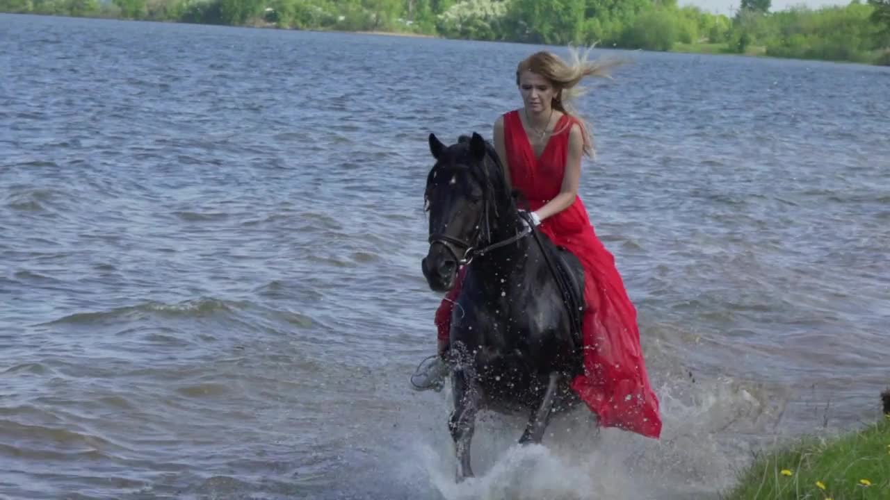 A beautiful blonde young sexy woman in a red dress riding a horse at a lake