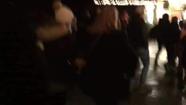 Guy walking through crowd at night trips black coat woman and she falls on face