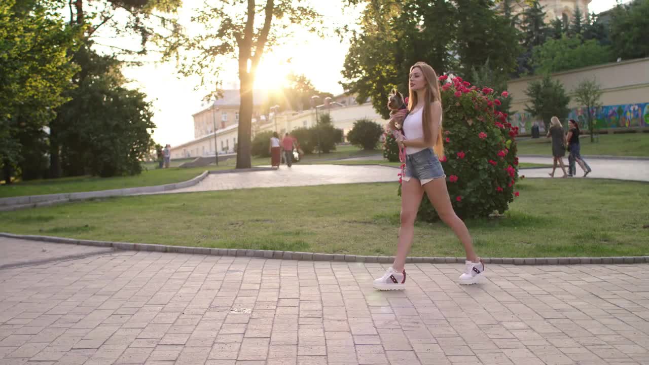 Beautiful Girl Walking With Her Small Dog In The City Park And Using Smartphone