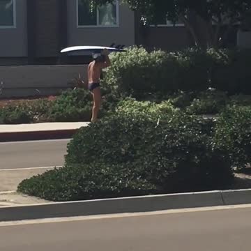 Guy in speedo carries surfboard on his head while barefoot