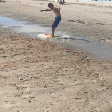 Blue shorts rides yellow skim board on beach and falls down