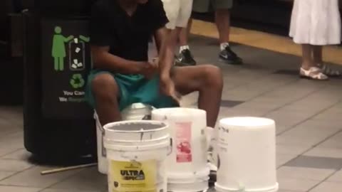 Guy in black shirt plays drums on buckets