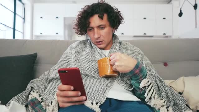 Young man having a bad cold and sitting on the sofa. He is drinking tea and using his smart phone.