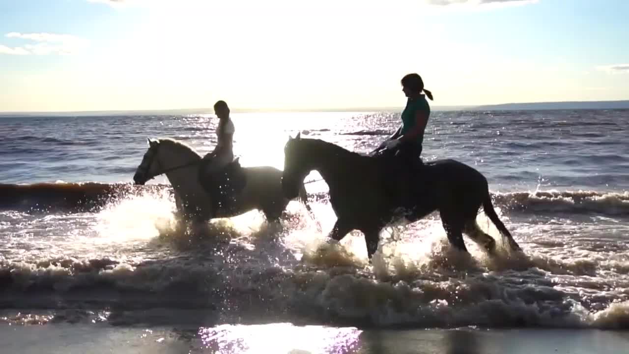 Two women ride on horse at river beach in water sunset light