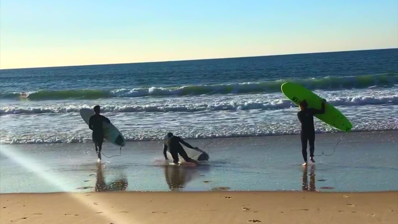 Three guys running to water with surfboard middle guy trips and falls into sand beach