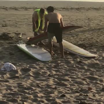 Two shirtless guys stack three surfboards and carries them off beach
