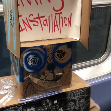 Cardboard box man sits on a subway train, "living installation"