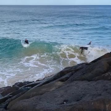 Wet suit guy falls off white surfboard into giant rocks