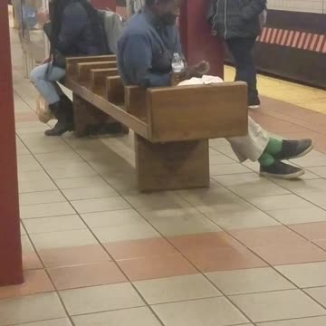 Homeless man smokes a cigarette and drinks out of water bottle on subway station bench
