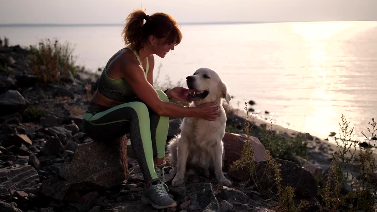 Woman stroking her dog on a shoreline at sunset