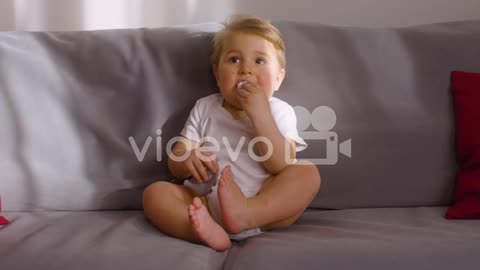 Front View Of A Baby Sitting On Sofa In Living Room At Home While Bitting A Toy Animal