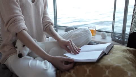 girl with a puppy reading a book sitting by the window with a lake view