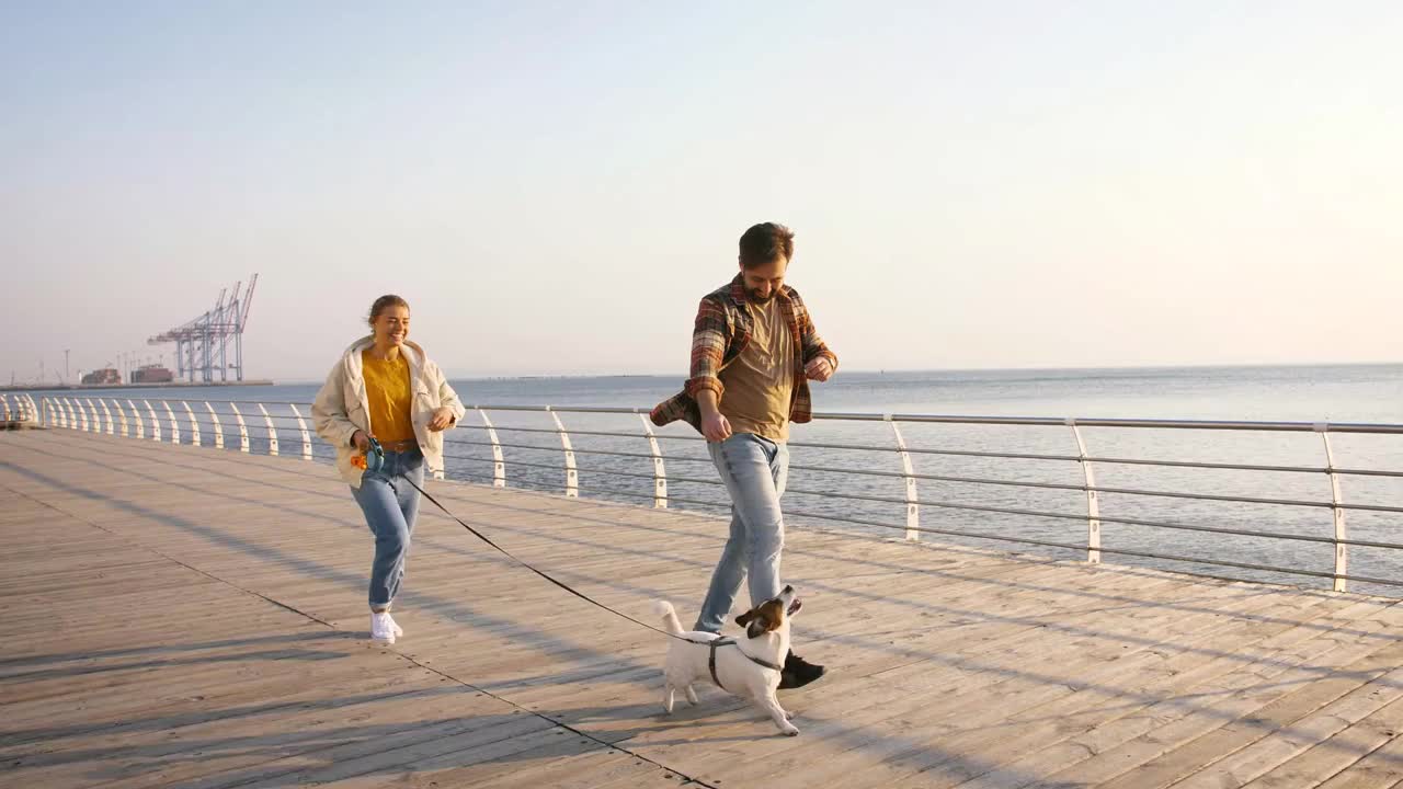 Cheerful young couple runnung and playing with cute Jack Russel terrier dog outdoors near the sea