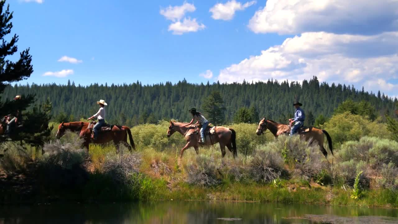 Group of people ride horses along mountainside trail and water