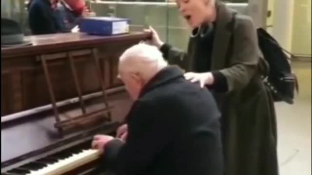 The old man is playing the piano, and the passing girls are singing along with it.