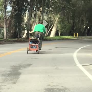 Green guy rides bicycle with wagon attached behind him