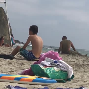 Old shirtless man puts stick in ear at beach plays with sand