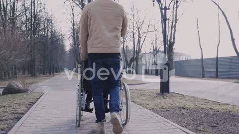 Rear View Of A Man Taking Her Disabled Friend In Wheelchair On A Walk Around The City