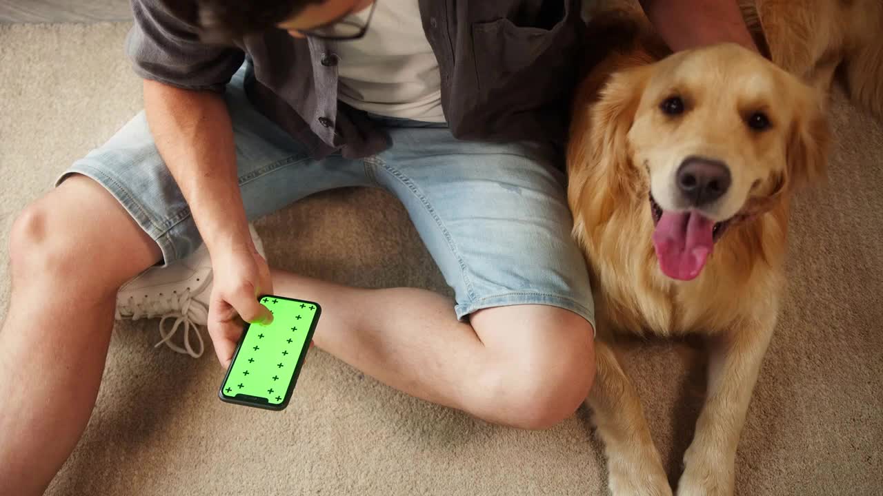 Young man in glasses petting golden retriever sitting on floor in living room