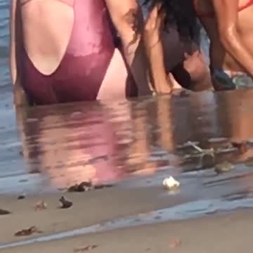 Guy records two girls sitting on beach one has bad tan lines